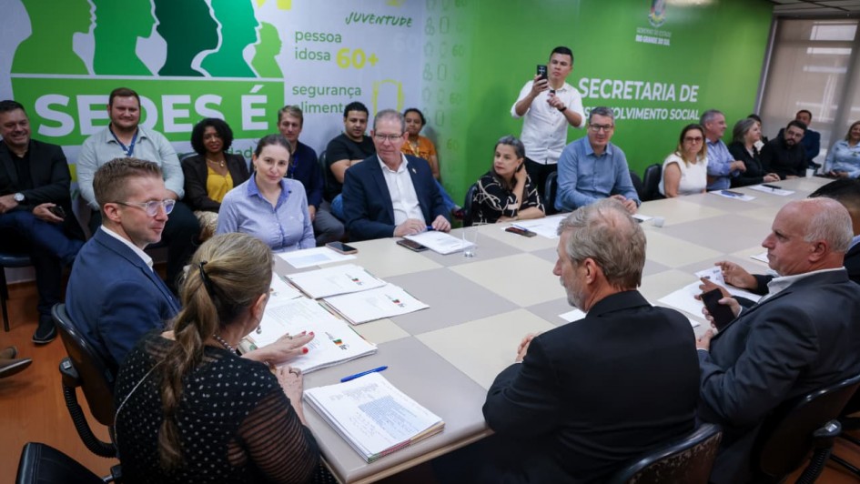 Foto de uma sala de reunião,  em que aparecem mais de 20 pessoas sentadas. Elas vestem trajes formais. Enquanto algumas conversam, outras observam. 