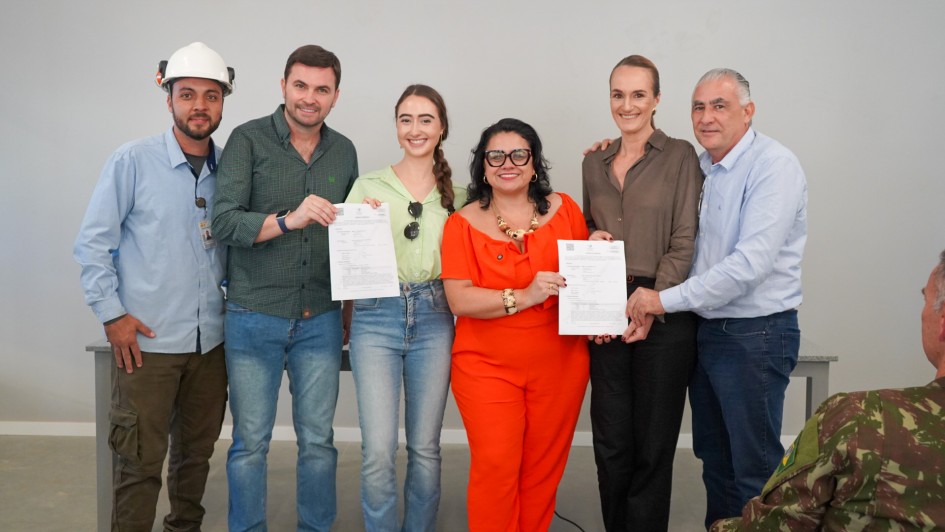 A imagem mostra seis pessoas em pé, posando para a foto, segurando documentos assinados. 