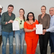 A imagem mostra seis pessoas em pé, posando para a foto, segurando documentos assinados. 