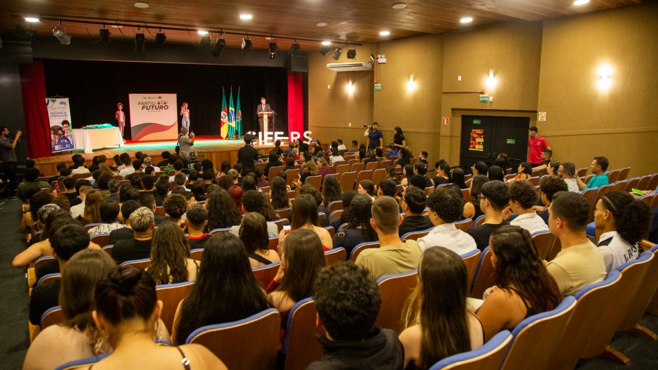 A fotografia mostra uma vista de um auditório lotado, capturada da parte de trás, olhando em direção ao palco.

Público: O auditório está cheio com uma grande plateia de jovens, sentados em fileiras de cadeiras estofadas marrons com detalhes em azul. As cabeças das pessoas no primeiro plano estão mais visíveis, e a maioria está olhando para o palco.

Palco: O palco, no fundo, tem um grande pano de fundo vermelho escuro e cortinas laterais pretas. No centro do palco, há uma mesa forrada com uma toalha branca e bandeiras do Brasil e do Rio Grande do Sul. Um homem está em pé atrás de um púlpito, discursando (provavelmente a cerimônia de formatura). À esquerda do palco, há um banner vertical e um pequeno grupo de pessoas.

Ambiente: O ambiente tem paredes laterais em tom de bege/marrom e um teto de madeira. Há luminárias embutidas no teto e arandelas nas paredes. Placas de saída de emergência são visíveis na parede lateral direita, onde também há duas portas escuras. O chão do auditório é inclinado (formato de anfiteatro).