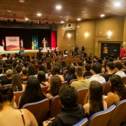 A fotografia mostra uma vista de um auditório lotado, capturada da parte de trás, olhando em direção ao palco.

Público: O auditório está cheio com uma grande plateia de jovens, sentados em fileiras de cadeiras estofadas marrons com detalhes em azul. As cabeças das pessoas no primeiro plano estão mais visíveis, e a maioria está olhando para o palco.

Palco: O palco, no fundo, tem um grande pano de fundo vermelho escuro e cortinas laterais pretas. No centro do palco, há uma mesa forrada com uma toalha branca e bandeiras do Brasil e do Rio Grande do Sul. Um homem está em pé atrás de um púlpito, discursando (provavelmente a cerimônia de formatura). À esquerda do palco, há um banner vertical e um pequeno grupo de pessoas.

Ambiente: O ambiente tem paredes laterais em tom de bege/marrom e um teto de madeira. Há luminárias embutidas no teto e arandelas nas paredes. Placas de saída de emergência são visíveis na parede lateral direita, onde também há duas portas escuras. O chão do auditório é inclinado (formato de anfiteatro).