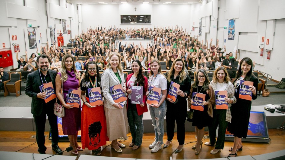 A foto mostra um auditório lotado, com estudantes e participantes levantando as mãos ao fundo. Na parte frontal do palco, um grupo de mulheres e homens posa para a foto segurando álbuns de figurinhas da Lei Maria da Penha. Todos estão alinhados em uma fileira, sorrindo para a câmera. O espaço é amplo, bem iluminado, com cadeiras ocupadas ao fundo e estrutura de auditório tradicional.