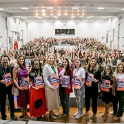 A foto mostra um auditório lotado, com estudantes e participantes levantando as mãos ao fundo. Na parte frontal do palco, um grupo de mulheres e homens posa para a foto segurando álbuns de figurinhas da Lei Maria da Penha. Todos estão alinhados em uma fileira, sorrindo para a câmera. O espaço é amplo, bem iluminado, com cadeiras ocupadas ao fundo e estrutura de auditório tradicional.