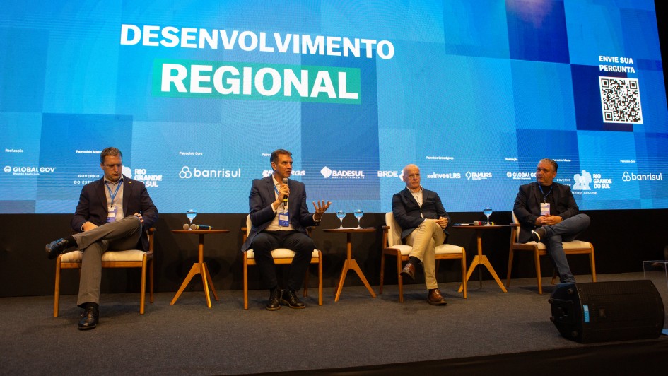 Foto de palco de cor escura com telão azul ao fundo. Quatro homens aparecem sentados. Um deles fala ao microfone.