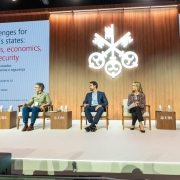 A imagem mostra um palco com cinco pessoas sentadas em poltronas. Uma delas fala ao microfone, enquanto as demais prestam atenção. Ao fundo, há um painel com o título da apresentação: "Challenges for Brazil's states: politics, economics, and security". 