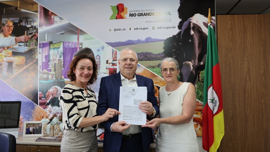 A imagem mostra três pessoas, duas mulheres e um homem, em um escritório, posando para uma foto enquanto seguram um documento.

Pessoas:

À esquerda, uma mulher de cabelo castanho cacheado, vestindo uma blusa estampada em preto e branco e calças claras.

No centro, um homem de óculos e terno azul, segurando um documento de cor branca com as duas mãos. Ele parece ser a figura central da foto.

À direita, uma mulher com cabelo grisalho e óculos, vestindo um vestido branco sem mangas.

Contexto e Fundo:

A cena ocorre em um escritório.

Na parede atrás das pessoas, há um grande painel ou banner promocional. O painel apresenta o logotipo do Governo do Estado do Rio Grande do Sul - Secretaria de Desenvolvimento Rural (SDR) e imagens relacionadas à vida rural e agronegócio (pessoas, animais, produtos, como queijo). Também são visíveis endereços de redes sociais e websites do governo.

À direita, próximo à mulher de branco, há uma bandeira do Rio Grande do Sul (verde, vermelho e amarelo) hasteada.

Parte de uma mesa de escritório e um monitor de computador são visíveis no canto esquerdo inferior.

Tema Geral: A foto parece registrar um encontro oficial, possivelmente relacionado à entrega de um documento ou acordo (mencionado no nome do arquivo como "Consulta Popular - São José do Sul") no contexto da Secretaria de Desenvolvimento Rural do Rio Grande do Sul.