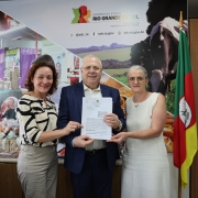 A imagem mostra três pessoas, duas mulheres e um homem, em um escritório, posando para uma foto enquanto seguram um documento.

Pessoas:

À esquerda, uma mulher de cabelo castanho cacheado, vestindo uma blusa estampada em preto e branco e calças claras.

No centro, um homem de óculos e terno azul, segurando um documento de cor branca com as duas mãos. Ele parece ser a figura central da foto.

À direita, uma mulher com cabelo grisalho e óculos, vestindo um vestido branco sem mangas.

Contexto e Fundo:

A cena ocorre em um escritório.

Na parede atrás das pessoas, há um grande painel ou banner promocional. O painel apresenta o logotipo do Governo do Estado do Rio Grande do Sul - Secretaria de Desenvolvimento Rural (SDR) e imagens relacionadas à vida rural e agronegócio (pessoas, animais, produtos, como queijo). Também são visíveis endereços de redes sociais e websites do governo.

À direita, próximo à mulher de branco, há uma bandeira do Rio Grande do Sul (verde, vermelho e amarelo) hasteada.

Parte de uma mesa de escritório e um monitor de computador são visíveis no canto esquerdo inferior.

Tema Geral: A foto parece registrar um encontro oficial, possivelmente relacionado à entrega de um documento ou acordo (mencionado no nome do arquivo como "Consulta Popular - São José do Sul") no contexto da Secretaria de Desenvolvimento Rural do Rio Grande do Sul.