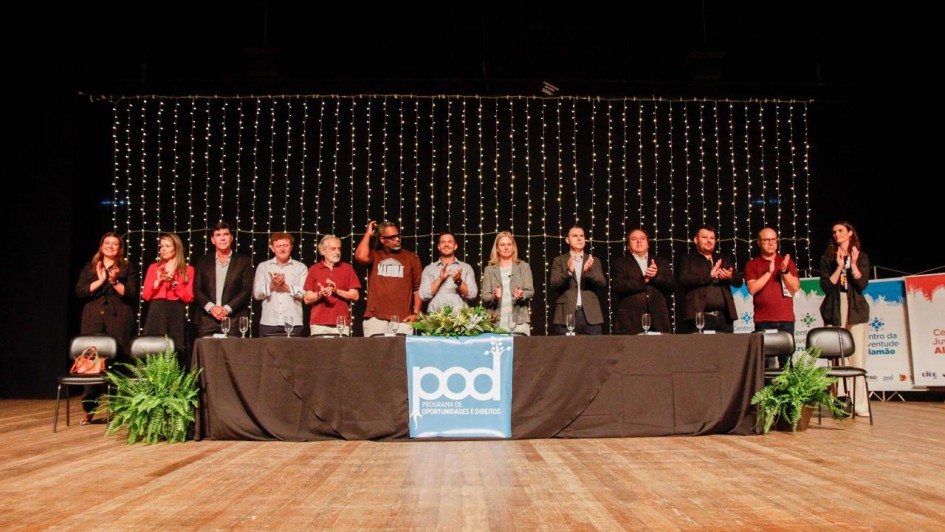 A imagem mostra uma solenidade em um palco amplo, decorado com luzes ao fundo, onde uma longa mesa coberta por uma toalha preta foi posicionada. Sobre ela, há um banner azul com o logotipo do Programa de Oportunidades e Direitos (POD). Em pé atrás da mesa, uma fileira de autoridades e convidados aplaude enquanto olha para o público. Há plantas ornamentais nas extremidades do palco. 