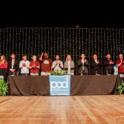 A imagem mostra uma solenidade em um palco amplo, decorado com luzes ao fundo, onde uma longa mesa coberta por uma toalha preta foi posicionada. Sobre ela, há um banner azul com o logotipo do Programa de Oportunidades e Direitos (POD). Em pé atrás da mesa, uma fileira de autoridades e convidados aplaude enquanto olha para o público. Há plantas ornamentais nas extremidades do palco. 