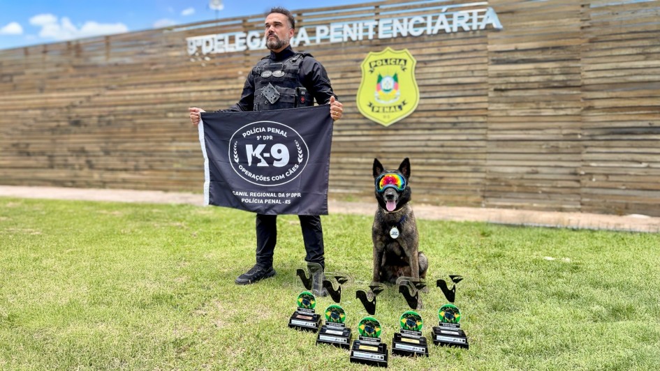 A imagem mostra um policial penal e um c&atilde;o de trabalho, em um ambiente externo, celebrando uma conquista com trof&eacute;us.

Pessoas e Animais:

Policial Penal: Em p&eacute; no lado esquerdo, um homem barbudo, vestindo uniforme t&aacute;tico preto (colete e cal&ccedil;as), olhando para a frente. Ele segura um estandarte/bandeira preto.

C&atilde;o K-9: Sentado no lado direito, est&aacute; um c&atilde;o de trabalho (possivelmente um Pastor Belga Malinois ou similar, com pelagem escura e tigrada). O c&atilde;o usa &oacute;culos de prote&ccedil;&atilde;o (goggles) nas cores neon (laranja, amarelo e azul) e est&aacute; com a boca aberta, parecendo estar ofegante e animado.

Bandeira (Estandarte): O policial segura uma bandeira preta com um emblema circular branco no centro. O texto vis&iacute;vel na bandeira &eacute;:

POL&Iacute;CIA PENAL e 9&ordf; DPR (na parte superior).

K-9 (grande, no centro).

OPERA&Ccedil;&Otilde;ES COM C&Atilde;ES (abaixo do K-9).

CANIL REGIONAL DA 9&ordf; DPR POL&Iacute;CIA PENAL - RS (na parte inferior).

Trof&eacute;us: Em primeiro plano, no gramado, h&aacute; uma fileira de cinco trof&eacute;us id&ecirc;nticos, iguais aos que aparecem na imagem anterior, simbolizando a conquista.

Fundo:

Eles est&atilde;o em um gramado verde, sob luz natural de um dia ensolarado, com c&eacute;u azul e algumas nuvens.

Atr&aacute;s deles, h&aacute; uma cerca ou parede feita de ripas de madeira horizontais.

No topo desta estrutura de madeira, h&aacute; uma placa com letras grandes e brancas que diz: 9&ordf; DELEGACIA PENITENCI&Aacute;RIA e, abaixo, POL&Iacute;CIA PENAL.

&Agrave; direita da placa, h&aacute; um bras&atilde;o ou distintivo da POL&Iacute;CIA PENAL em cores verdes, amarelas e vermelhas.

Tema Geral: A imagem celebra uma vit&oacute;ria ou conquista de um c&atilde;o de trabalho (K-9) da Pol&iacute;cia Penal do Rio Grande do Sul (9&ordf; DPR), destacando o c&atilde;o, seu condutor, os trof&eacute;us e a identifica&ccedil;&atilde;o da institui&ccedil;&atilde;o.