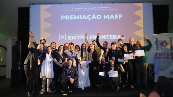 A imagem registra um grande grupo de pessoas no palco, celebrando um evento de premiação.

Pessoas e Ação: Cerca de 15 a 20 pessoas (homens e mulheres) estão no palco, posando juntas. A maioria está vestindo roupas casuais a semi-formais e acessórios (como crachás de evento). Muitas pessoas estão levantando os braços e as mãos em sinal de celebração e entusiasmo. Algumas seguram certificados ou documentos.

Fundo (Tela de Projeção): A tela de projeção, que domina o fundo, exibe o tema do evento.

No topo, lê-se: "5º Mercado Audiovisual Entre Fronteras" e, em uma linha menor, o slogan: "Onde as fronteiras se diluem, as histórias se encontram".

No centro, em letras grandes e em negrito: "PREMIAÇÃO MAEF" (sendo MAEF a sigla do Mercado Audiovisual Entre Fronteras).

Abaixo, há o logotipo estilizado do "5º MERCADO AUDIOVISUAL ENTRE FRONTERAS".

Ambiente: A cena se passa em um ambiente interno, provavelmente um auditório ou salão de eventos. O palco é elevado e há caixas de som visíveis nas laterais. A iluminação é focada no palco.

Outros Detalhes: No lado direito, parte de um banner promocional do evento "ENTRE FRONTERAS" é visível, repetindo a identificação do evento. No canto inferior direito, o crédito do fotógrafo "crédito Sérgio Rosa" é mencionado no nome do arquivo.

Tema Geral: A imagem é um retrato de grupo no palco durante a cerimônia de premiação do 5º Mercado Audiovisual Entre Fronteras, mostrando a alegria e celebração dos participantes ou vencedores.