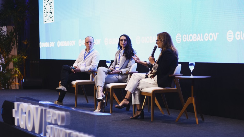 Foto de palco com três pessoas sentadas: duas mulheres e um homem. A mulher da direita fala ao microfone.
