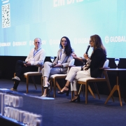 Foto de palco com três pessoas sentadas: duas mulheres e um homem. A mulher da direita fala ao microfone.