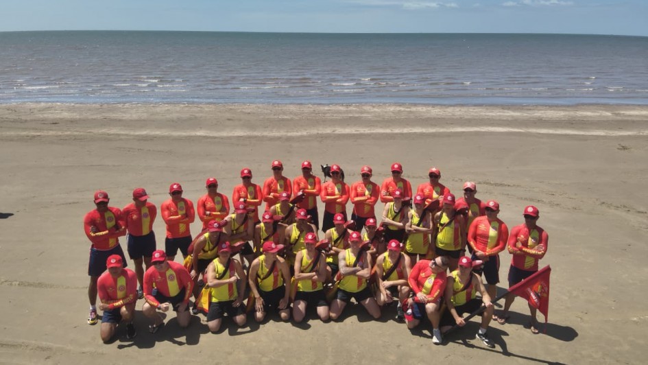 A foto mostra um grande grupo de guarda-vidas reunidos para uma foto coletiva na praia. Todos vestem uniformes vermelhos e amarelos, com bonés vermelhos. Ao fundo, há o mar e um horizonte limpo sob céu azul claro.