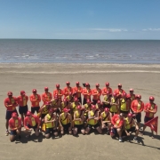 A foto mostra um grande grupo de guarda-vidas reunidos para uma foto coletiva na praia. Todos vestem uniformes vermelhos e amarelos, com bonés vermelhos. Ao fundo, há o mar e um horizonte limpo sob céu azul claro.