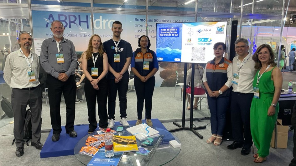 Foto posada de oito pessoas em ambiente fechado, numa sala de exposição no simpósio. A pessoas estão de pé e usam crachá, e encontram-se próximas a uma mesa baixa com livros e folhetos e a uma TV ao fundo.