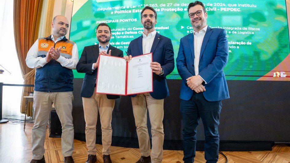 Quatro homens posam para foto em um salão nobre, em frente a um painel digital sobre o Sistema Estadual de Proteção e Defesa Civil. Ao centro, o governador Eduardo Leite segura e exibe um documento oficial aberto, acompanhado pelo vice-governador Gabriel Souza, que aparece imediatamente à esquerda dele (na foto). Os outros dois integrantes, aplaudem.