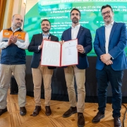Quatro homens posam para foto em um salão nobre, em frente a um painel digital sobre o Sistema Estadual de Proteção e Defesa Civil. Ao centro, o governador Eduardo Leite segura e exibe um documento oficial aberto, acompanhado pelo vice-governador Gabriel Souza, que aparece imediatamente à esquerda dele (na foto). Os outros dois integrantes, aplaudem.