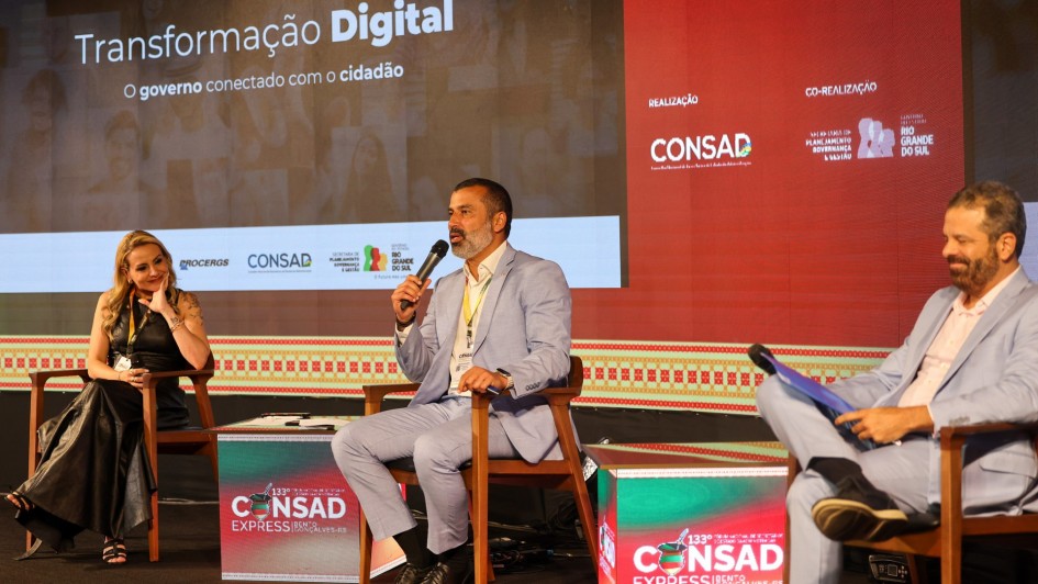 Três painelistas estão sentados em cadeiras, com cubos coloridos entre eles, com o nome do evento. O mediador, ao centro, está falando ao microfone. À esquerda dele na foto, há uma painelista mulher, loira, com vestido marrom, que o olha atentamente. Do outro lado, um homem, de terno claro, sorri olhando para o papel. 