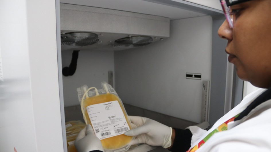 Uma foto de um homem em pé em um salão de conferências (possivelmente Gabriel Souza), no canto esquerdo, discursando em um evento de lançamento de concessão. Ele veste um blazer escuro, camisa branca e calças cáqui claras, e gesticula em direção a uma tela de LED grande e escura que ocupa a parede à esquerda.

A tela exibe texto em letras amarelas e brancas, com a palavra "escola" e a frase "é agora" parcialmente visíveis, indicando o tema da apresentação.

No centro e à direita, há uma audiência de cerca de 15 pessoas, sentadas em cadeiras e poltronas, assistindo à apresentação. Alguns fotógrafos e cinegrafistas estão de pé na parte de trás da sala. Em primeiro plano, uma pequena mesa redonda preta contém copos d'água e crachás. O ambiente é moderno, com iluminação de trilhos e grandes janelas horizontais que oferecem vista para uma área externa com árvores e edifícios. A cena sugere o lançamento da concessão do serviço de bilhetagem eletrônica do transporte metropolitano.