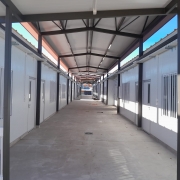Uma foto de um corredor coberto (passarela ou área de circulação) em um complexo de edifícios modulares (contêineres ou salas pré-fabricadas), possivelmente uma escola. O corredor é longo e estreito, com simetria acentuada.

As paredes laterais dos prédios são brancas. As janelas, também brancas, possuem grades de proteção verticais e horizontais. Portas brancas de entrada para as salas estão visíveis ao longo de ambos os lados do corredor.

A estrutura da cobertura é sustentada por vigas escuras (pretas ou marrom-escuras) de metal, formando um telhado inclinado. O piso do corredor é de cimento. A iluminação é fornecida por luminárias brancas de LED fixadas na parte inferior do telhado. A luz natural entra pela abertura superior da cobertura, e no fundo, é visível uma área de pátio e construções adjacentes.