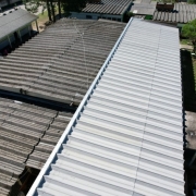 Uma foto aérea com ângulo inclinado, mostrando o telhado de um complexo de edifícios, possivelmente uma escola. A imagem destaca um grande telhado novo, de metal cor cinza-claro e formato ondulado, que cobre uma longa estrutura retangular no centro da foto.

Este telhado novo contrasta com os telhados antigos ao redor. Os telhados adjacentes são de amianto (ou material similar), de cor cinza escura e com marcas de tempo e sujeira. A luz do sol incide sobre o telhado novo, refletindo a cor clara.

Ao redor das estruturas, há pequenas áreas de pátio cimentado e árvores. A imagem sugere a revitalização ou reforma da cobertura de uma parte do complexo escolar.
