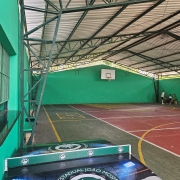 Uma foto de uma quadra poliesportiva coberta (possivelmente de um colégio estadual) com foco em uma mesa de tênis de mesa (pingue-pongue) em primeiro plano.

Em primeiro plano:

A mesa de tênis de mesa tem uma superfície de jogo verde-escura e um design no corpo principal (a lateral da mesa).

O design da mesa apresenta o nome "COLÉGIO ESTADUAL JOÃO MOSMANN" em arco, circundando um emblema ou logotipo com as iniciais "C E J M" e o mapa do Rio Grande do Sul.

A iluminação azul e verde é projetada artificialmente no design da mesa.

A quadra:

A quadra é coberta por um telhado de metal sustentado por uma estrutura metálica de treliças escuras (verde-escuras ou pretas).

O piso da quadra é vermelho e verde (com divisões amarelas) e exibe marcações para esportes (o círculo central é visível).

A parede de fundo é de cor verde-água/turquesa vibrante e apresenta uma tabela de basquete branca com aros.

No canto direito, dois jovens estão sentados em um banco ou área de descanso.

A estrutura lateral da quadra (à esquerda) é gradeada com barras verticais. A foto sugere uma área de lazer e esportes de uma instituição de ensino.