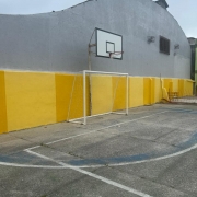 
Uma foto da fachada de um prédio escolar sob um céu azul-claro com nuvens. A fachada do edifício de dois andares (vista lateral em ângulo aberto) é de cor cinza-claro.

O que se destaca é a aplicação de painéis retangulares coloridos entre as janelas de cada andar, em uma sequência vibrante:

Um painel em vermelho.

Um painel em verde.

Um painel em amarelo.

Um painel em verde-musgo.

Um painel em laranja/vermelho queimado.

Um painel em verde-oliva.

As janelas são múltiplas janelas-guilhotina (tipo basculante) com grades de metal finas (quadriculadas) e escuras. O telhado é cinza escuro. À esquerda da construção, há árvores frondosas, e na base do prédio, em primeiro plano, há um canteiro de terra e um pequeno caminho cimentado. A revitalização utiliza cores para dar um aspecto moderno e alegre à escola.
