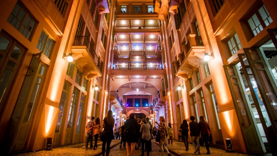A imagem mostra a Casa de Cultura Mario Quintana (CCMQ) a noite. Iluminada em tons quentes, entre as paredes rosadas, pessoas não identificadas caminham na Travessa dos Cataventos, que divide as alas da CCMQ.