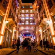 A imagem mostra a Casa de Cultura Mario Quintana (CCMQ) a noite. Iluminada em tons quentes, entre as paredes rosadas, pessoas não identificadas caminham na Travessa dos Cataventos, que divide as alas da CCMQ.