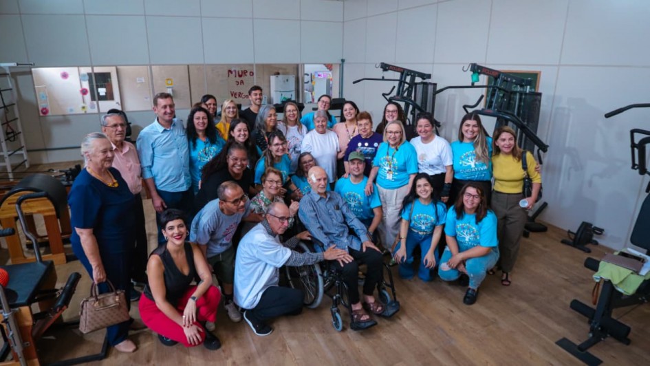 Foto de diversas pessoas em ambiente fechado, muitas com camisa azul da instituição. Elas posam pra foto e sorriem. Há um idoso numa cadeira de rodas no centro.
