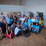 Foto de diversas pessoas em ambiente fechado, muitas com camisa azul da instituição. Elas posam pra foto e sorriem. Há um idoso numa cadeira de rodas no centro.
