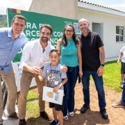 O governador e uma família posam para a foto em frente a uma casa entregue. Eles sorriem e uma criança segura a chave da moradia.