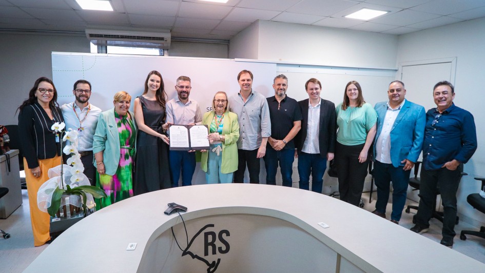 Um grupo de doze pessoas está em pé posando para uma foto. Ao centro, a titular da SES exibe um documento assinado. 