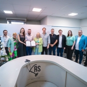 Um grupo de doze pessoas está em pé posando para uma foto. Ao centro, a titular da SES exibe um documento assinado. 