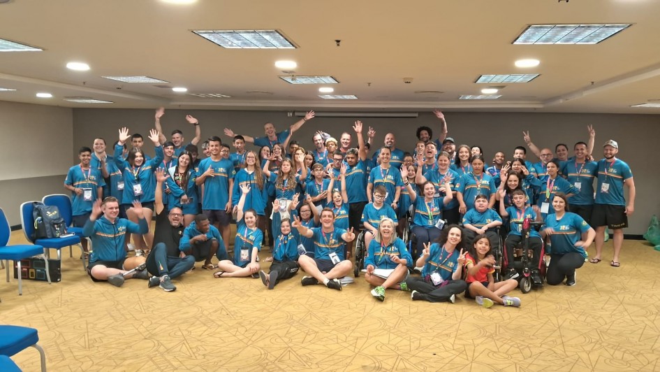 Uma foto externa e panorâmica de uma grande delegação gaúcha (cerca de 50 a 60 pessoas, de todas as idades) reunida em uma sala de conferências ou salão de hotel. O grupo parece ser a delegação para as Paralimpíadas de 2025.

As Pessoas:

A maioria está vestindo camisetas polo azuis-turquesa com detalhes amarelos nos ombros e no colarinho, que são os uniformes da delegação.

Os membros do grupo estão sentados no chão, agachados ou em pé, e muitos levantam os braços e acenam, sorrindo de forma entusiasmada.

Diversos atletas em cadeiras de rodas estão visíveis na primeira fileira e no centro da imagem.

O Ambiente:

A sala tem iluminação clara vinda de luminárias embutidas no teto em padrões retangulares.

O chão é acarpetado com um padrão geométrico em tons de amarelo e marrom-claro.

No canto esquerdo, algumas cadeiras azuis de plástico estão empilhadas.

A cena transmite um clima de união, celebração e entusiasmo.