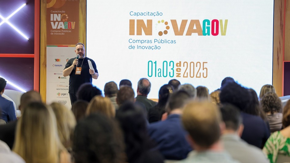 Uma foto de uma profissional da saúde, no Hemocentro do Rio Grande do Sul (**HEMORGS**), ao lado de uma outra mulher, inspecionando um refrigerador para armazenamento de sangue e hemoderivados.

À esquerda, está um **ultracongelador/refrigerador laboratorial** com a porta de cor branca aberta, revelando seu interior com bandejas metálicas. Próximo ao refrigerador, há equipamentos semelhantes nas cores cinza-claro e azul, identificados por marcas comerciais (não totalmente legíveis).

Duas mulheres estão voltadas para o refrigerador:
1.  **À esquerda, a mulher de cabelo grisalho**, vestindo uma blusa branca e um cardigã preto, inclina-se para a frente, olhando para o interior do equipamento.
2.  **À direita, a mulher de cabelo louro-claro na altura dos ombros**, veste um colete de cor bege com o texto **"MINISTERIO DA SAÚDE"** (parcialmente visível) em letras escuras nas costas. Ela parece estar lendo um papel preso a um dos equipamentos ao lado.

O ambiente é um laboratório, com piso de cor clara e paredes de azulejo branco com linhas escuras. No fundo, há uma porta entreaberta com moldura de madeira marrom. No lado direito, mais equipamentos de armazenamento brancos são visíveis, com etiquetas de aviso de segurança.