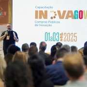 Uma foto de uma profissional da saúde, no Hemocentro do Rio Grande do Sul (**HEMORGS**), ao lado de uma outra mulher, inspecionando um refrigerador para armazenamento de sangue e hemoderivados.

À esquerda, está um **ultracongelador/refrigerador laboratorial** com a porta de cor branca aberta, revelando seu interior com bandejas metálicas. Próximo ao refrigerador, há equipamentos semelhantes nas cores cinza-claro e azul, identificados por marcas comerciais (não totalmente legíveis).

Duas mulheres estão voltadas para o refrigerador:
1.  **À esquerda, a mulher de cabelo grisalho**, vestindo uma blusa branca e um cardigã preto, inclina-se para a frente, olhando para o interior do equipamento.
2.  **À direita, a mulher de cabelo louro-claro na altura dos ombros**, veste um colete de cor bege com o texto **"MINISTERIO DA SAÚDE"** (parcialmente visível) em letras escuras nas costas. Ela parece estar lendo um papel preso a um dos equipamentos ao lado.

O ambiente é um laboratório, com piso de cor clara e paredes de azulejo branco com linhas escuras. No fundo, há uma porta entreaberta com moldura de madeira marrom. No lado direito, mais equipamentos de armazenamento brancos são visíveis, com etiquetas de aviso de segurança.