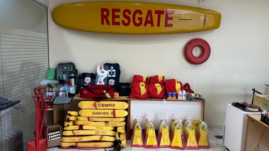 A imagem mostra uma sala com diversos equipamentos de salvamento utilizados por guarda-vidas. Na parede, há uma prancha de resgate amarela com a palavra “RESGATE” em vermelho. Ao lado, está pendido um flutuador circular vermelho. Sobre uma mesa, encontram-se mochilas vermelhas, bolsas de primeiros socorros, cilindros de oxigênio, colar cervical, protetor solar e outros itens médicos. No chão, empilhados, há vários flutuadores amarelos com a inscrição “CBMRS – GUARDA-VIDAS”, além de pares de nadadeiras também nas cores amarela e vermelha embaladas em plástico. À esquerda, aparecem pás e outros materiais de apoio.