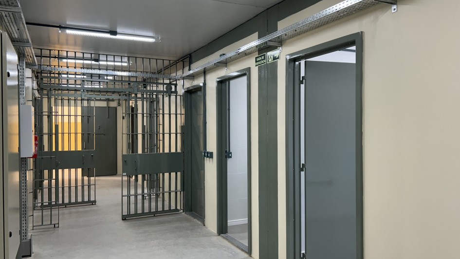 A imagem é uma fotografia de um corredor interno da Casa de Custódia da Polícia Civil em Porto Alegre, focada em um ambiente que combina elementos de segurança institucional com uma infraestrutura nova ou reformada.

Elementos da Cena:
Estrutura de Segurança (Esquerda e Fundo):

No lado esquerdo e no fundo, há grades metálicas escuras (celas ou portões de segurança), típicas de áreas de custódia.

Um portão de grade está aberto, revelando um interior com paredes amarelas/alaranjadas escuras.

Corredor e Paredes (Direita):

O corredor principal possui um piso de cimento cinza claro e paredes de cor bege claro/amarelado.

Há portas fechadas de cor cinza-escura ao longo da parede direita, indicando possíveis salas ou celas individuais. Uma das portas está ligeiramente aberta, revelando um interior de cor clara, possivelmente um banheiro.

As portas têm batentes e detalhes verticais de cor cinza-escura, contrastando com a parede.

Infraestrutura e Iluminação:

O teto é baixo e claro, equipado com luminárias de LED tubulares (fluorescentes) que fornecem iluminação branca e intensa.

Na parte superior das paredes e teto, há calhas metálicas (eletrocalhas) expostas, indicando a passagem de cabos elétricos ou de dados, o que sugere uma instalação moderna ou recente.

Há também uma placa de sinalização de saída de emergência ("SAÍDA") verde e branca acima de uma das portas.

Conclusão Visual:
A foto representa a área interna de custódia, destacando a combinação de elementos de segurança (grades) com uma infraestrutura moderna (iluminação e cabeamento exposto), possivelmente resultado de uma obra de construção ou reforma.