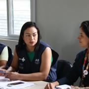 A imagem mostra três mulheres sentadas lado a lado, em uma mesa de trabalho, vestindo coletes com inscrições do SUS e SAES. Elas conversam entre si e seguram canetas e papéis. 
