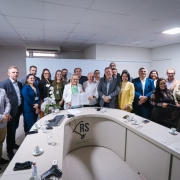 Um grupo numeroso de pessoas está em pé, reunido para a foto, atrás de uma mesa em formato de U. Ao centro, a secretária da Saúde segura o documento assinado. 