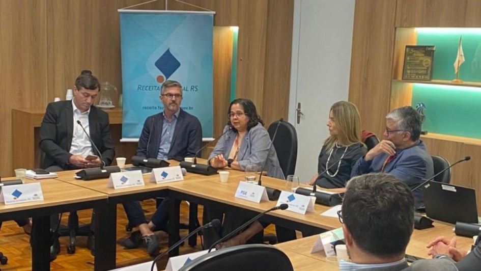 Foto mostra equipe reunida ao redor de uma mesa em sala com fundo de madeira.