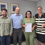 A imagem mostra quatro pessoas em pé, reunidas para a foto. A titular da Sema segura e exibe um documento assinado. 