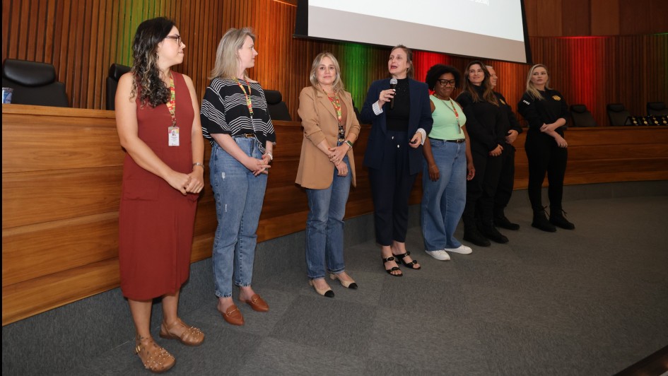 Foto meio de lado de 7 mulheres e 1 homem à frente de um auditório. Eles estão de pé e, ao centro, uma mulher fala ao microfone.