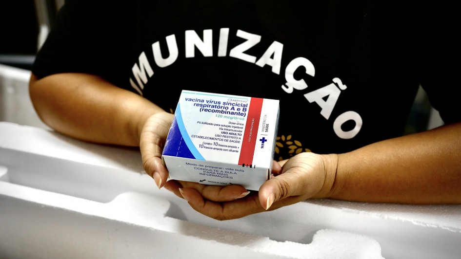 Foto das mãos de uma mulher segurando a caixa da vacina, de cor predominantemente branca. Ela usa uma camiseta preta em que se lê Imunização.