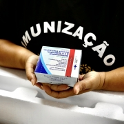 Foto das mãos de uma mulher segurando a caixa da vacina, de cor predominantemente branca. Ela usa uma camiseta preta em que se lê Imunização.