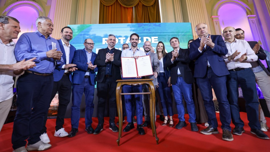 A foto mostra um grupo de aproximadamente 15 pessoas reunidas em um palco com carpete vermelho, dentro de um salão amplo com colunas claras e iluminação em tons de roxo e amarelo. Ao centro, o governador Eduardo Leite, segurando um documento aberto com ambas as mãos, posicionado sobre uma mesa pequena e estreita. As pessoas ao redor batem palmas. Atrás do grupo, há um grande painel digital exibindo cores em azul e verde.