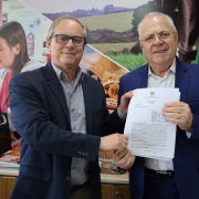 A imagem mostra dois homens segurando um documento assinado e posando para a foto. 