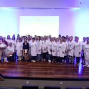 Um grande grupo de pessoas está reunido em um auditório, posicionado sobre um palco de madeira. A maioria veste jalecos brancos e crachás verdes. O grupo ocupa quase toda a largura do palco e está organizado em várias fileiras, olhando para a câmera. Em frente ao grupo há uma mulher vestindo roupas escuras e uma saia estampada, posicionada ao centro. Ao fundo, um telão iluminado em azul exibe informações sobre o evento, embora o texto não seja totalmente legível. A sala tem luzes frias que criam um brilho azul nas paredes.